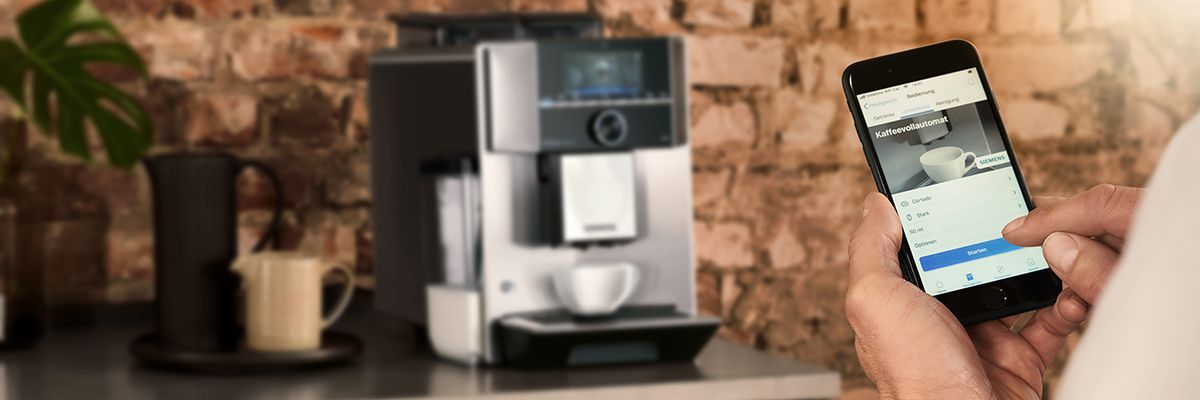 Eine Person steuert mit dem Smartphone einen Kaffeevollautomaten, im Hintergrund steht die Kaffeemaschine auf einer Theke.